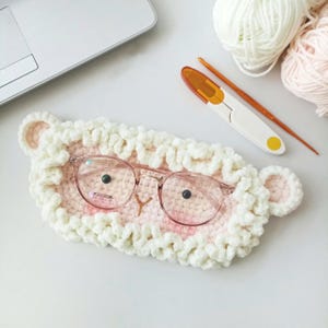 Puede incluir: Un estuche para gafas de crochet blanco y rosa hecho a mano con forma de oveja, con gafas redondas con montura rosa en su interior. El estuche tiene una textura esponjosa y orejas de oveja. También se ven unas tijeras, un ganchillo y lana.