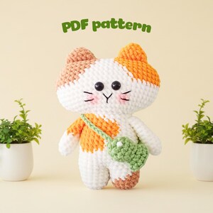 Puede incluir: Muñeca de gato de ganchillo con detalles blancos, naranjas y marrones, con una bolsa verde. La muñeca tiene ojos negros y mejillas rosadas. El texto "PDF pattern" está encima de la muñeca. Dos pequeñas plantas en macetas flanquean la muñeca.