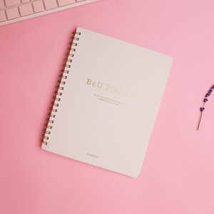 May include: A white spiral bound notebook with the text "Be U Planner" in gold lettering on a pink background. A single lavender stem is on the right side of the notebook.