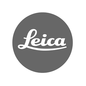 Op de afbeelding: Een rond logo met een effen donkergrijze achtergrond en het woord Leica in een wit, gestileerd cursief lettertype, horizontaal gecentreerd in het midden van de cirkel. De tekst is strak, goed leesbaar en vetgedrukt.