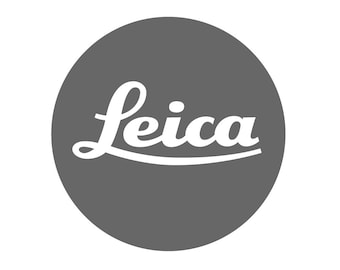 May include: A circular logo featuring a solid dark grey background with the word Leica written in a white, stylised, cursive script font centred horizontally across the middle of the circle. The text is clean, legible, and bold.