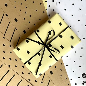 Könnte beinhalten: Nahaufnahme eines Geschenks, das in gelbes Papier mit schwarzen Quadraten und einem schwarzen Band eingewickelt ist. Das Geschenk ist von Blättern braunen Geschenkpapiers mit schwarzen geometrischen Mustern umgeben.