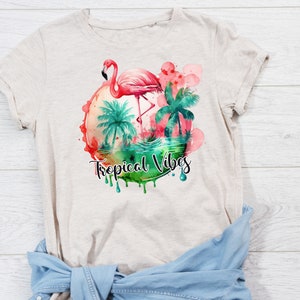 May include: A beige t-shirt with a watercolor design of a pink flamingo standing in a tropical scene with palm trees. The design is surrounded by a circle of pink and green watercolor and the text "Tropical Vibes" is written below the circle.
