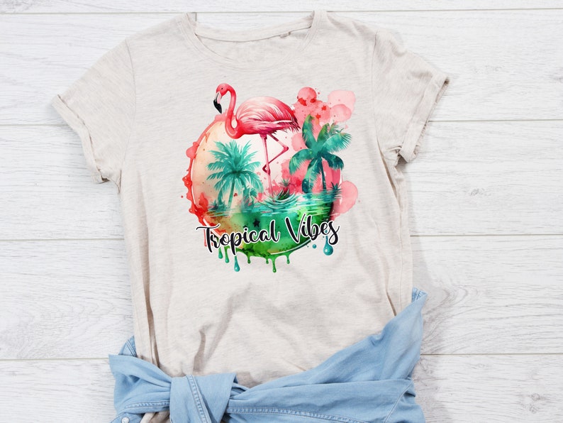 May include: A beige t-shirt with a watercolor design of a pink flamingo standing in a tropical scene with palm trees. The design is surrounded by a circle of pink and green watercolor and the text "Tropical Vibes" is written below the circle.