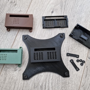 May include: Assortment of 3D printed plastic cases in black, brown, and green. The cases have various shapes and sizes, with some featuring a grid pattern. There are also several black plastic standoffs and washers.