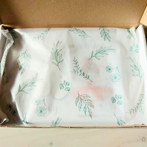Pu&ograve; includere: Una scatola di spedizione rettangolare in cartone con coperchio aperto, contenente carta velina bianca stampata con un motivo ripetuto di foglie botaniche, steli e piccoli fiori simili a denti di leone verdi, che coprono parzialmente un oggetto.