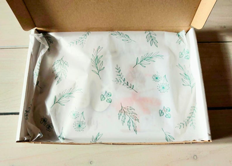 Pu&ograve; includere: Una scatola di spedizione rettangolare in cartone con coperchio aperto, contenente carta velina bianca stampata con un motivo ripetuto di foglie botaniche, steli e piccoli fiori simili a denti di leone verdi, che coprono parzialmente un oggetto.