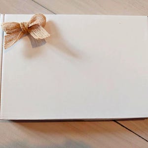 Pu&ograve; includere: Una scatola postale rettangolare in cartone bianco con un fiocco decorativo in nastro di iuta color marrone chiaro fissato nell'angolo in alto a sinistra. La scatola ha una superficie pulita e liscia e un design pieghevole standard e funzionale.