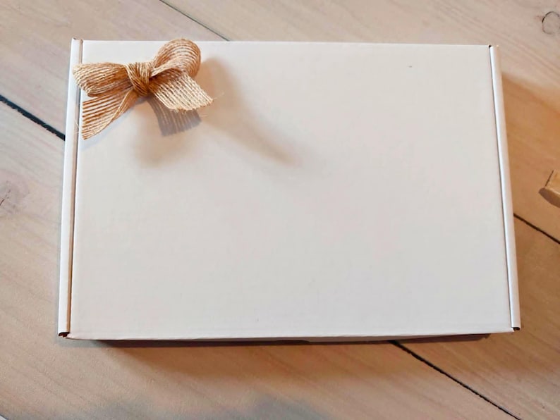 Pu&ograve; includere: Una scatola postale rettangolare in cartone bianco con un fiocco decorativo in nastro di iuta color marrone chiaro fissato nell'angolo in alto a sinistra. La scatola ha una superficie pulita e liscia e un design pieghevole standard e funzionale.