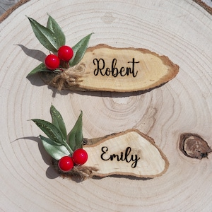 May include: Two wooden place cards, one with "Robert" and the other with "Emily" in black script. Each is adorned with red berries, green leaves, and twine. They rest on a light wood surface, ideal for a rustic event.
