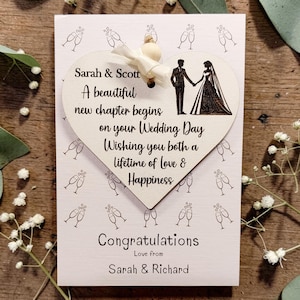 May include: A wedding card with a heart-shaped tag featuring a bride and groom silhouette, with text that reads "Sarah & Scott" and "Congratulations". The card is decorated with champagne glasses and floral accents.