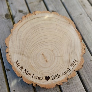 May include: A circular wooden slice with a natural bark edge, engraved with the text "Mr & Mrs Jones", a heart symbol, and "28th April 2024". The wood has a light colour. Suitable for wedding decorations or personalised gifts.