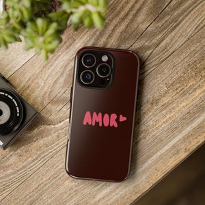 May include: A dark brown phone case with the word "AMOR" in pink script. The case has a glossy finish and a camera cutout. The phone case is resting on a wooden surface.