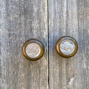 May include: Two antique cabinet knobs with a brass finish. One knob features a profile of a Native American, the other a buffalo. The knobs are made from vintage five-cent coins.