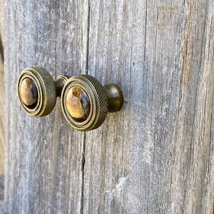 May include: Two antique brass cabinet knobs with a ridged edge and a central oval of brown and gold stone. The knobs are mounted on a weathered wooden surface, suggesting a vintage or rustic aesthetic.