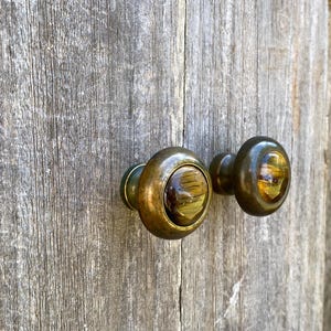 May include: Two antique brass door knobs with a tiger's eye stone center. The knobs are round with a dark, aged finish, mounted on a weathered wooden surface. The tiger's eye stone has a mix of brown and gold tones.