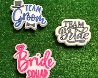 May include: Three white and colorful charms with text for a wedding. One charm says "Team Groom" with a top hat and bow tie. Another says "Team Bride". The third charm says "Bride Squad" with a diamond ring.