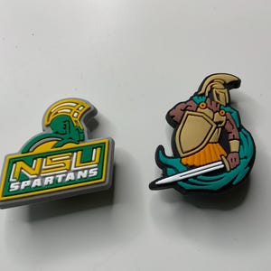 Op de afbeelding: Twee kleurrijke schoenbedels. Eén is een rechthoekig ontwerp met de tekst "NSU SPARTANS" in groen en geel. De andere is een cartoon Spartaanse krijger met een zwaard, schild en helm.