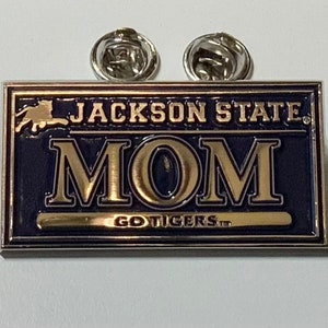 May include: A rectangular pin with the words "JACKSON STATE MOM" in gold lettering on a navy blue background. The pin also features the words "GO TIGERS" in gold. Two silver pin backs are attached.