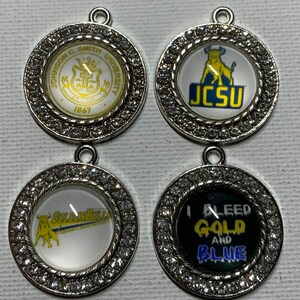 May include: Four silver charms with rhinestones around the edge. Each charm has a different design. The first charm has a gold crest with the text "Johnson C. Smith University 1867". The second charm has a yellow bull with the text "JCSU". The third charm has a yellow bull with the text "Golden Bulls". The fourth charm has a black background with the text "I Bleed Gold and Blue".