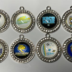 May include: Eight round silver charms with clear rhinestone borders. Each charm features a different design, including text and graphics related to HBCU and college themes. The charms are likely used for jewelry or accessories.