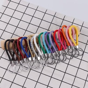 Puede incluir: Una colección de llaveros de cuero trenzado en varios colores: marrón, rojo, azul, plateado, blanco, verde, morado, rosa, naranja y amarillo. Cada llavero tiene un anillo de metal plateado.
