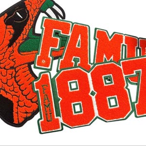 Könnte beinhalten: Ein orangefarbener und grüner Aufnäher mit einem Schlangenkopf und dem Text "FAMU 1887". Der Aufnäher hat einen schwarzen Rand und einen weißen Hintergrund. Der Text ist grün umrandet.