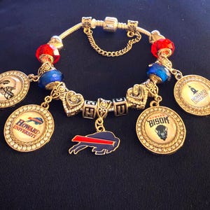 Op de afbeelding: Een zilverkleurige bedelarmband met rode en blauwe gefacetteerde kralen, hartvormige tussenstukjes en letterkralen die HU spellen. Het bevat ronde bedels met Howard University logo's, een footballhelm en een blauw met rode bizon-silhouetbedel.