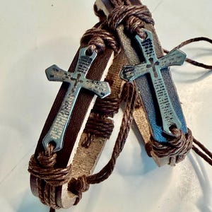 May include: Two brown leather bracelets, each featuring a metal cross pendant. The bracelets have a woven rope design and the crosses have etched details. The leather straps are tan and brown.