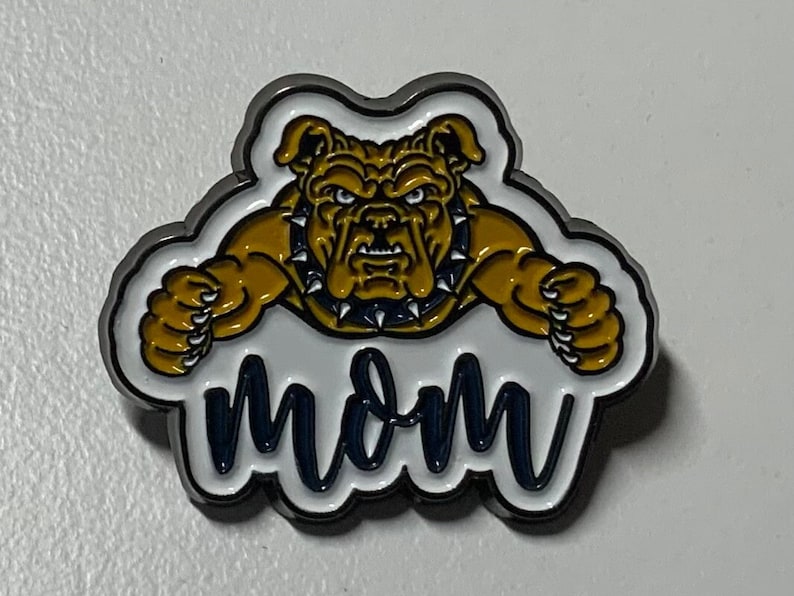 May include: A silver pin with a white background and a cartoon bulldog in yellow, blue, and black. The bulldog is wearing a black collar with spikes. The text "MOM" is written in blue on the white background.