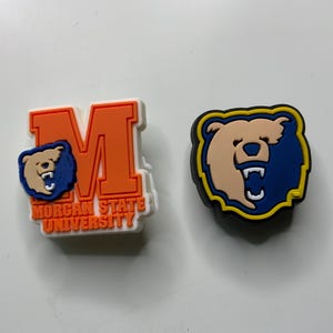 Op de afbeelding: Twee kleurrijke schoenbedels. Eén is oranje en wit met een blauw berenlogo en de tekst "MORGAN STATE UNIVERSITY". De andere is een berenlogo met een blauwe en gele rand.