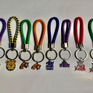 Puede incluir: Una colección de ocho llaveros con correas de cuero trenzado en varios colores, incluyendo morado, amarillo, verde, naranja, azul, rojo y plateado. Cada llavero presenta un dije metálico con diferentes diseños, como un bulldog, una serpiente y varios logotipos.