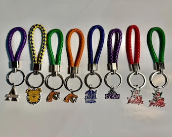 Puede incluir: Una colección de ocho llaveros con correas de cuero trenzado en varios colores, incluyendo morado, amarillo, verde, naranja, azul, rojo y plateado. Cada llavero presenta un dije metálico con diferentes diseños, como un bulldog, una serpiente y varios logotipos.