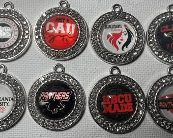 May include: Eight silver charms with rhinestones around the edges. Each charm features a different design, including a book, a panther, a woman's face, and the words "CAU", "Clark Atlanta University Alumni", "Panthers", and "HBCU Made".