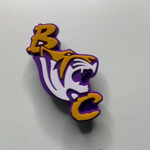 Könnte beinhalten: Ein lila, weißer und goldener Schuhanhänger mit einem Tigerkopf und den Buchstaben "BC". Der Tiger hat weiße Zähne und ein gold-weiß gestreiftes Gesicht. Die Buchstaben "BC" sind in Gold gehalten.