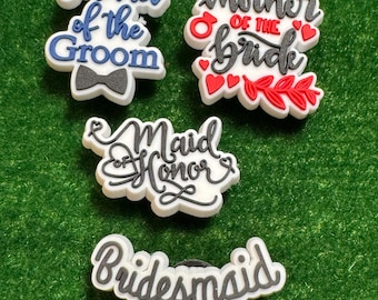 May include: A collection of white, blue, red, and black shoe charms with wedding-themed text. The charms include phrases like "Mother of the Groom," "Mother of the Bride," "Maid of Honor," and "Bridesmaid." The charms are on a green surface.