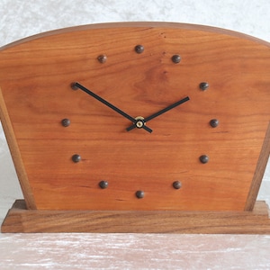 May include: A wooden clock with a curved top and a dark wood base. The clock face is made of light wood and has black hands and black metal accents.
