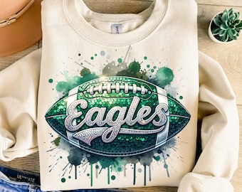 May include: Cream-colored sweatshirt featuring a green and silver football design with the word "Eagles". The football has a sequined texture and is set against a watercolor splash background in shades of green.