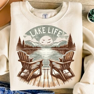 May include: Cream-colored sweatshirt with a lakeside graphic. The design features two wooden Adirondack chairs on a dock, a lake scene with mountains, trees, and the text "LAKE LIFE" in a vintage style.