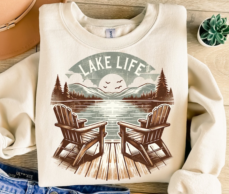 May include: Cream-colored sweatshirt with a lakeside graphic. The design features two wooden Adirondack chairs on a dock, a lake scene with mountains, trees, and the text "LAKE LIFE" in a vintage style.