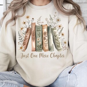 Op de afbeelding: Cr&egrave;mekleurige sweatshirt met een afbeelding van boeken en bloemen, en de tekst "Just One More Chapter". De boeken zijn in tinten bruin, groen en perzik, met witte madeliefjes en groen. Een comfortabel ontwerp voor boekenliefhebbers.