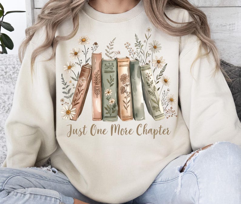 Op de afbeelding: Cr&egrave;mekleurige sweatshirt met een afbeelding van boeken en bloemen, en de tekst "Just One More Chapter". De boeken zijn in tinten bruin, groen en perzik, met witte madeliefjes en groen. Een comfortabel ontwerp voor boekenliefhebbers.