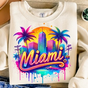 May include: A beige sweatshirt with a colorful graphic design of the Miami skyline, palm trees, and the word "Miami" in neon lettering. The design has a watercolor effect with dripping paint.