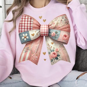 May include: A light pink sweatshirt featuring a large, decorative bow design. The bow is composed of various patterned fabrics, including gingham, floral, and striped patterns, in shades of pink, red, and blue. Gold heart accents are scattered around the bow.