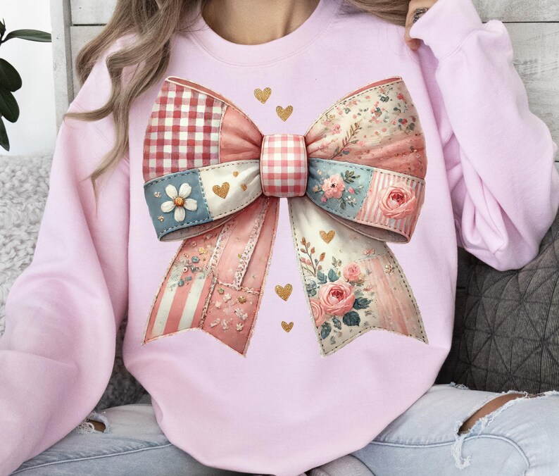 May include: A light pink sweatshirt featuring a large, decorative bow design. The bow is composed of various patterned fabrics, including gingham, floral, and striped patterns, in shades of pink, red, and blue. Gold heart accents are scattered around the bow.