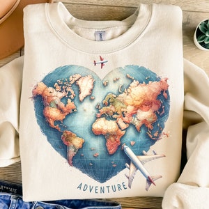 Może przedstawiać: Kremowa bluza z nadrukiem w kształcie serca z mapą świata i kontynentami w stylu akwareli. Słowo "ADVENTURE" jest wydrukowane pod mapą. Przedstawiono dwa samoloty.