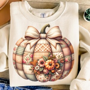 May include: Cream-colored sweatshirt featuring a large, detailed pumpkin design. The pumpkin is composed of patchwork patterns in shades of orange, pink, green, and cream, with a large bow and floral accents. A fall-themed graphic.