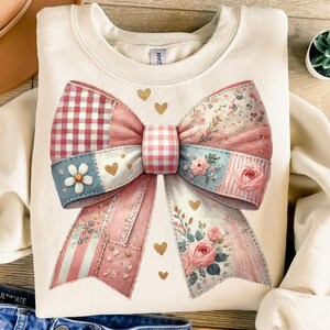 May include: Cream-colored sweatshirt featuring a large bow design. The bow is a patchwork of patterns, including gingham, floral prints, and stripes, in shades of pink, blue, and red. Gold heart accents are scattered around the bow.