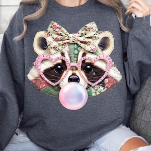 Könnte beinhalten: Ein dunkelgraues Sweatshirt mit einem skurrilen Waschbär-Design. Der Waschbär hat eine florale Schleife, herzförmige Brille und eine Kaugummiblase. Das Gesicht des Waschbären besteht aus Patchwork-Stoff.