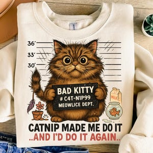 May include: Cream-colored sweatshirt featuring a brown tabby cat holding a "Bad Kitty" sign. The graphic includes a milk carton, a fishbowl, and the text "Catnip Made Me Do It...And I'd Do It Again." The design has a humorous theme.